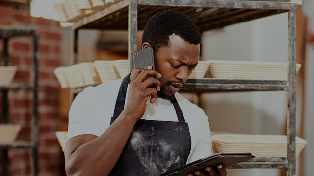 A man working in a bakery wearing an apron is on a mobile phone and looking at a tablet.